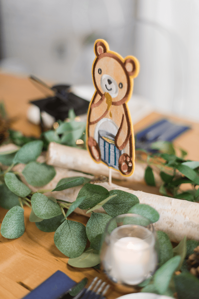 Woodland-themed baby boy shower centerpiece featuring a fabric bear eating honey, perched on a birch log with flowing greenery, rustic lanterns, and scattered pinecones for a charming forest-inspired display.