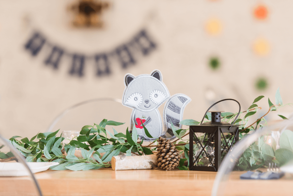 Woodland-themed baby boy shower centerpiece featuring a fabric raccoon perched on a birch log, surrounded by flowing greenery, lanterns, and pinecones for rustic, nature-inspired decor.