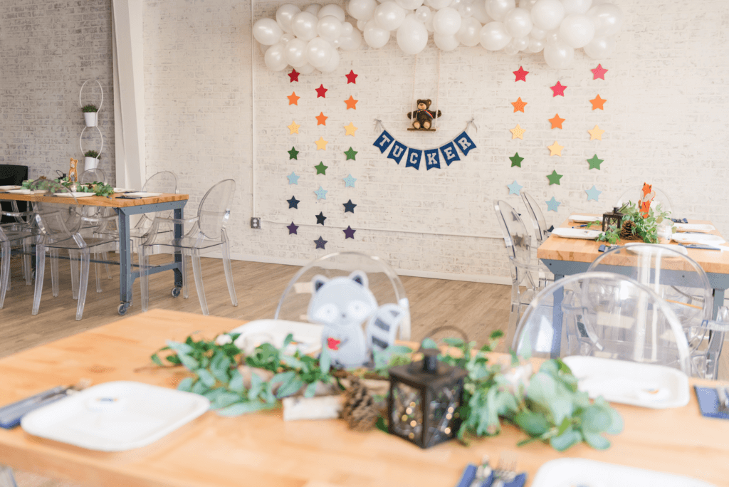 Woodland-themed baby boy shower decor featuring a white brick backdrop with white balloon clouds and rainbow star garland. A plush teddy bear sits on a wooden swing, creating a whimsical backdrop for a woodland-themed baby boy shower.