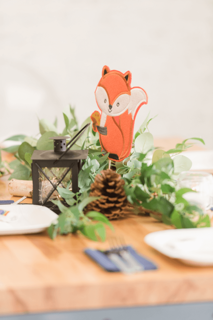 Woodland-themed baby shower centerpiece featuring a fabric squirrel perched on a birch log, surrounded by flowing greenery, lanterns with fairy lights, and pinecones.