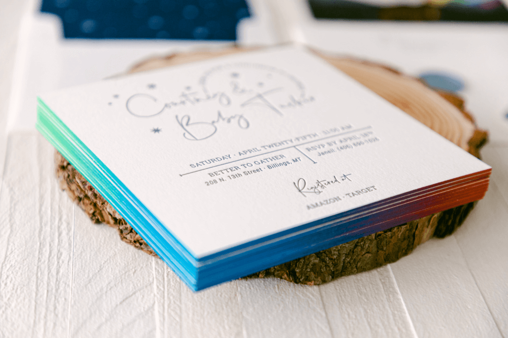 Woodland-themed baby boy shower letterpress invitation featuring rainbow hand-painted edges, placed on a rustic wood slice.