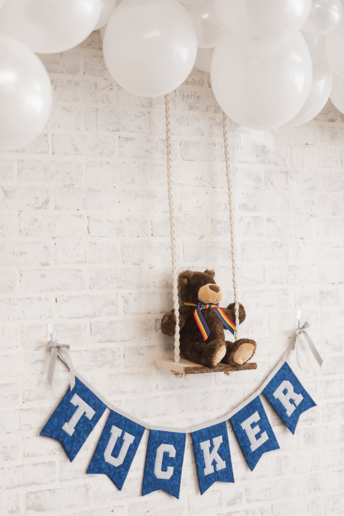 Closeup of a teddy bear sitting on a wooden swing with a white brick backdrop highlighting the fabric garland below the swing, displaying the baby boy's name.