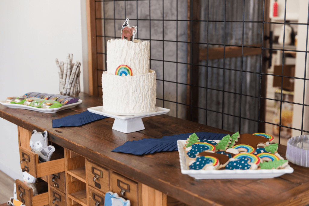 Woodland-themed baby boy shower dessert table displayed on a rustic wooden cart with whimsical woodland animals peeking out from the drawers.