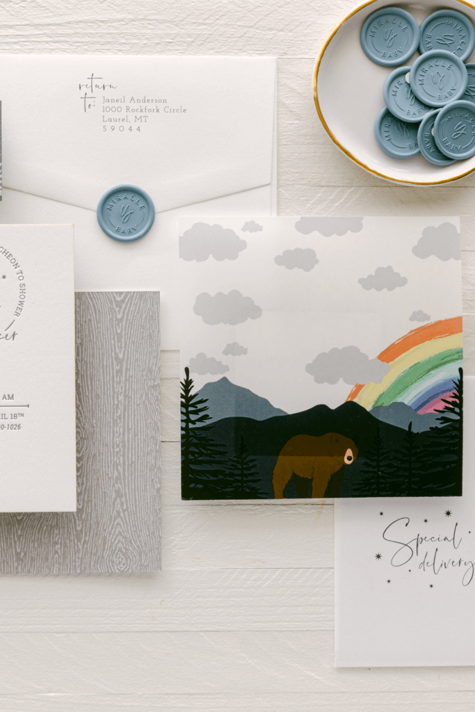 Woodland-themed baby boy shower invitation with custom illustrated vellum overlay featuring a rainbow, pine trees, and a bear, designed to reflect a nature-inspired celebration for a rainbow baby.