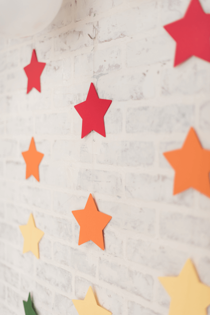 Closeup of rainbow star garland at a woodland-themed baby boy shower, set against a white brick backdrop. The colorful stars add a playful touch, complementing the natural woodland elements and symbolizing the joy of a rainbow baby.