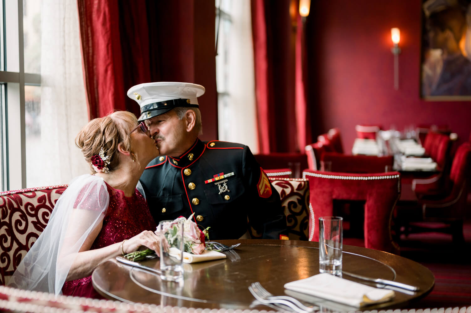 Celebrating 50 Years of Love: Our Parents' Golden Anniversary 29 The couple shares a kiss at a café table before the event, dressed in Marine Corps dress blues and a red gown.
