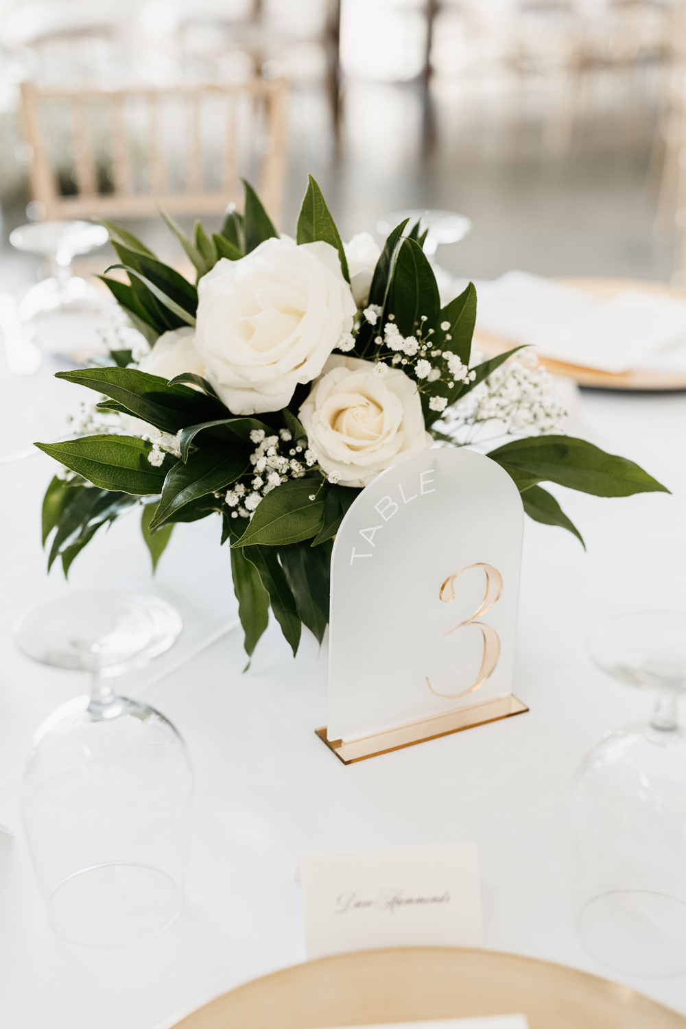 Black and White Wedding Invitations for a Classic Ashford Acres Inn Wedding 22 Table setting featuring a white floral arrangement with greenery, acrylic table number with gold lettering, and gold-accented place settings at a black and white wedding reception.