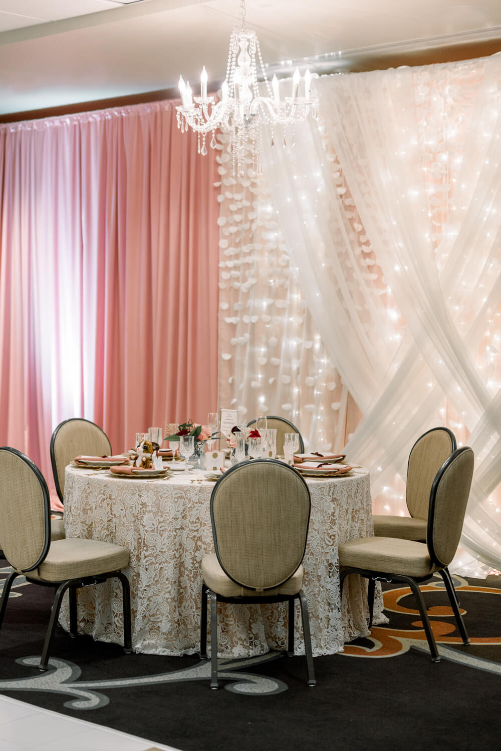 Celebrating 50 Years of Love: Our Parents' Golden Anniversary 7 Close-up of a Victorian-inspired table setting at a 50th wedding anniversary, featuring lace linens, blush napkins, crystal glassware, and a soft pink and ivory backdrop with twinkle lights and a chandelier