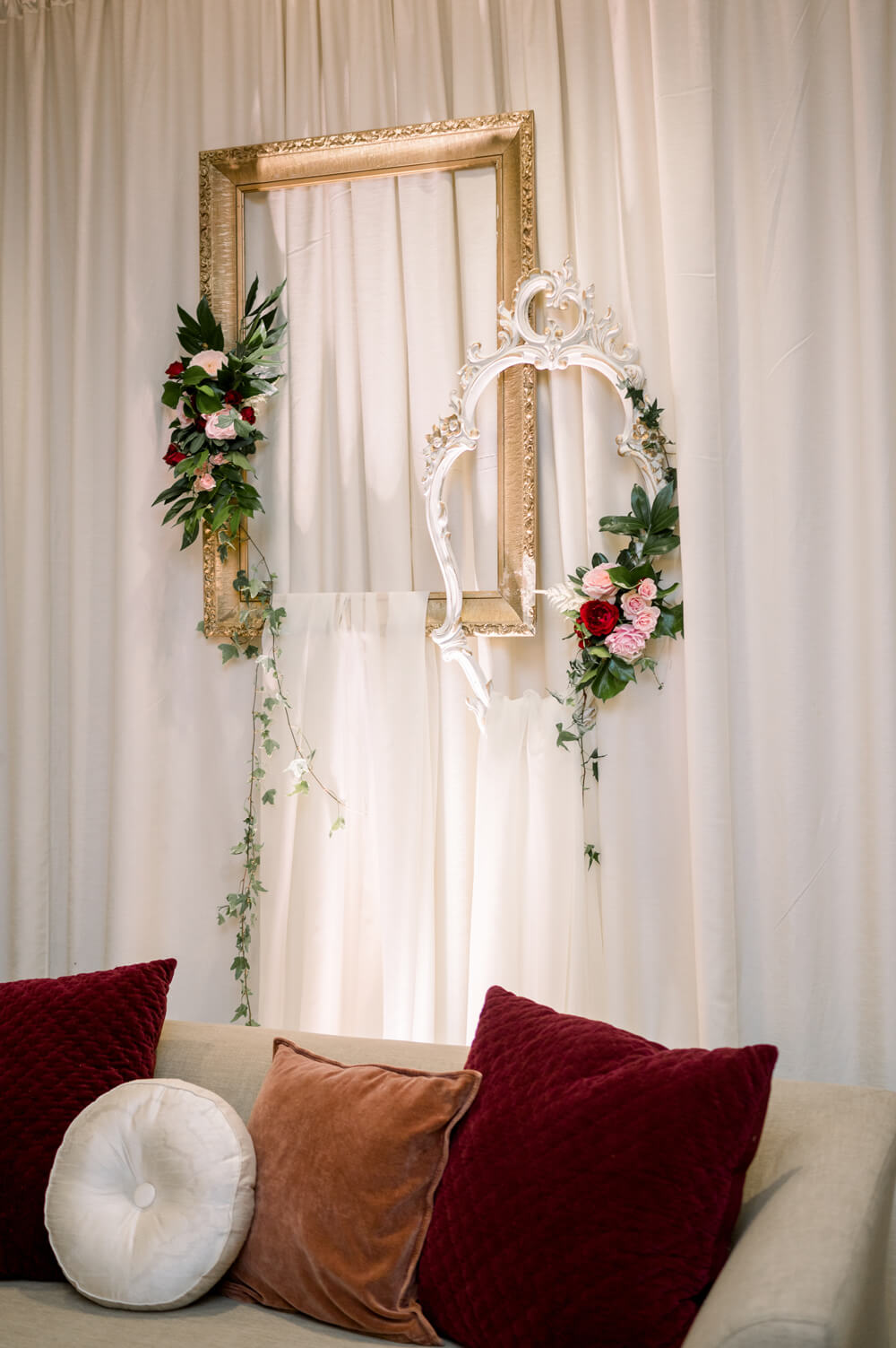 Celebrating 50 Years of Love: Our Parents' Golden Anniversary 11 Close-up of a Victorian-inspired lounge backdrop with layered ivory drapery, ornate gold and white frames, and burgundy and rust velvet pillows for a 50th anniversary celebration.