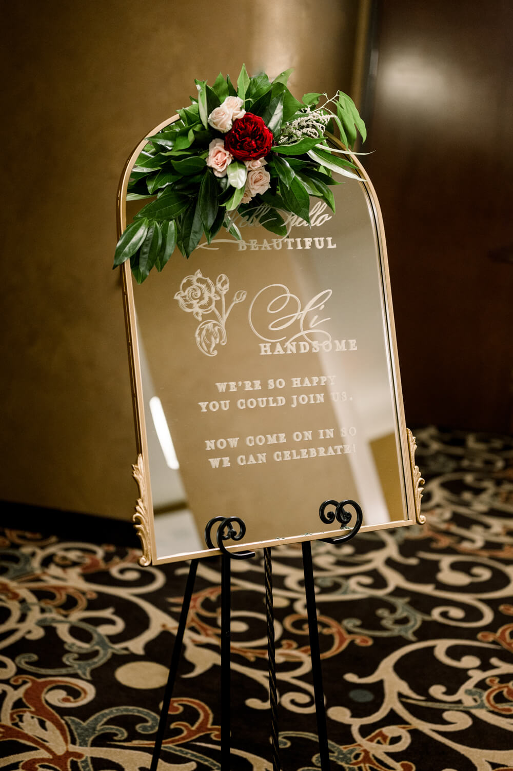 Celebrating 50 Years of Love: Our Parents' Golden Anniversary 17 Gold-framed mirror welcome sign adorned with fresh burgundy and blush florals, greeting guests to a golden 50th wedding anniversary celebration.