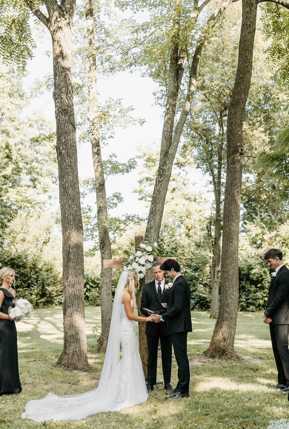 Black and White Wedding Invitations for a Classic Ashford Acres Inn Wedding 12 The bride and groom holding hands during their vows while standing in front of a flower-covered wooden cross, with trees and guests in view.