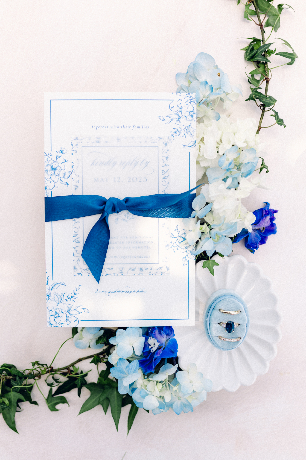 Flat lay of blue and white wedding invitation suite tied with silk ribbon, surrounded by florals and ring dish, showcasing refined stationery and soft garden-inspired styling.