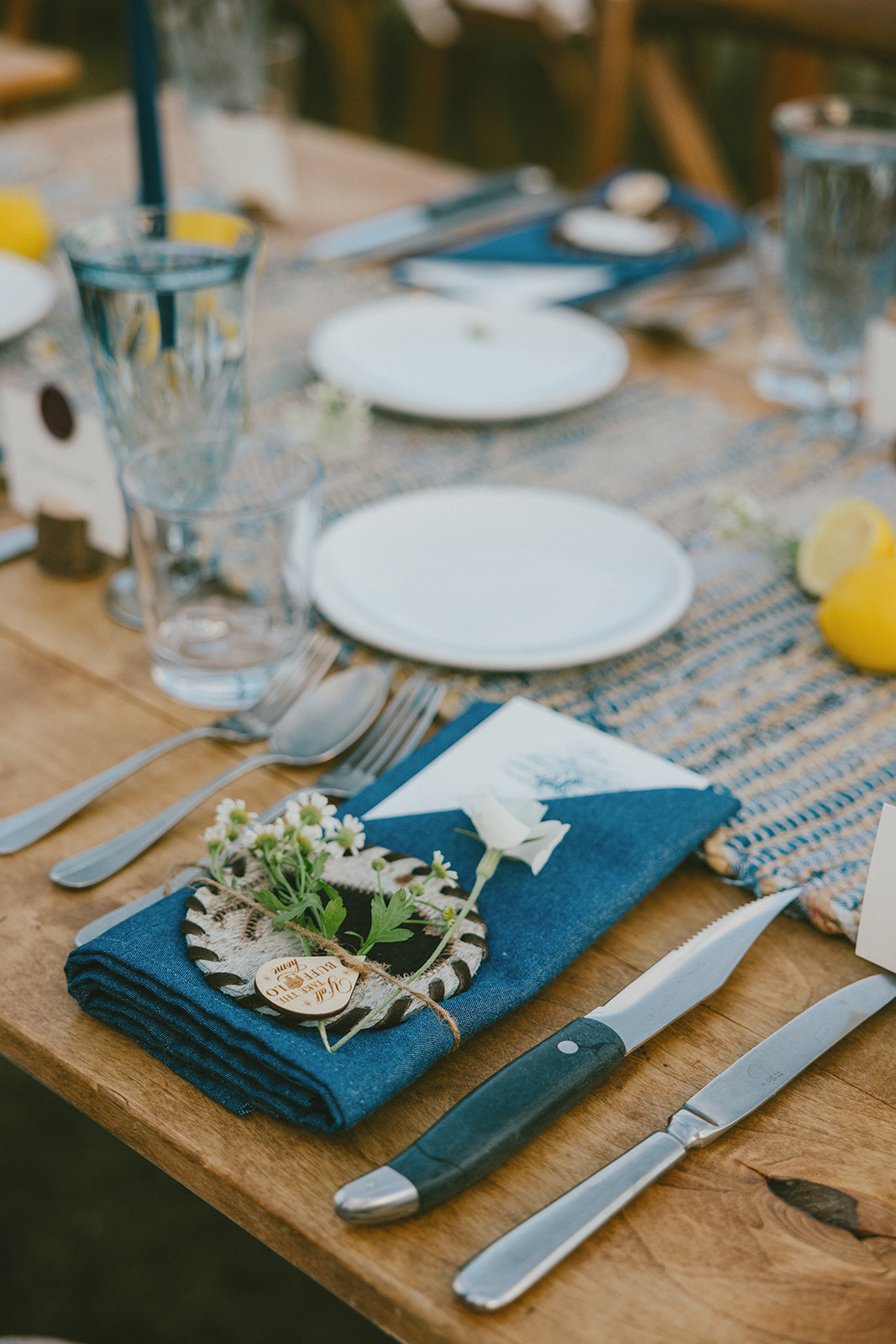 Rustic place setting with blue linen napkin, wood place card, cutlery, and glassware arranged on a wooden table, styled for an intimate outdoor wedding reception.