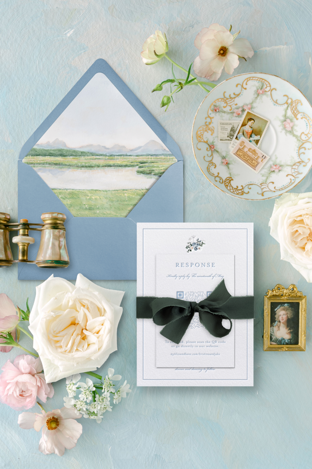 How to Choose a Stationery Designer When “Pretty” Isn’t Enough 4 Flat lay of a refined wedding invitation suite featuring a blue envelope with a hand-painted landscape liner, ribbon-tied RSVP card, floral accents, vintage stamps, and porcelain details on a soft blue background, styled to highlight thoughtful design and guest experience. | how to choose a stationery designer