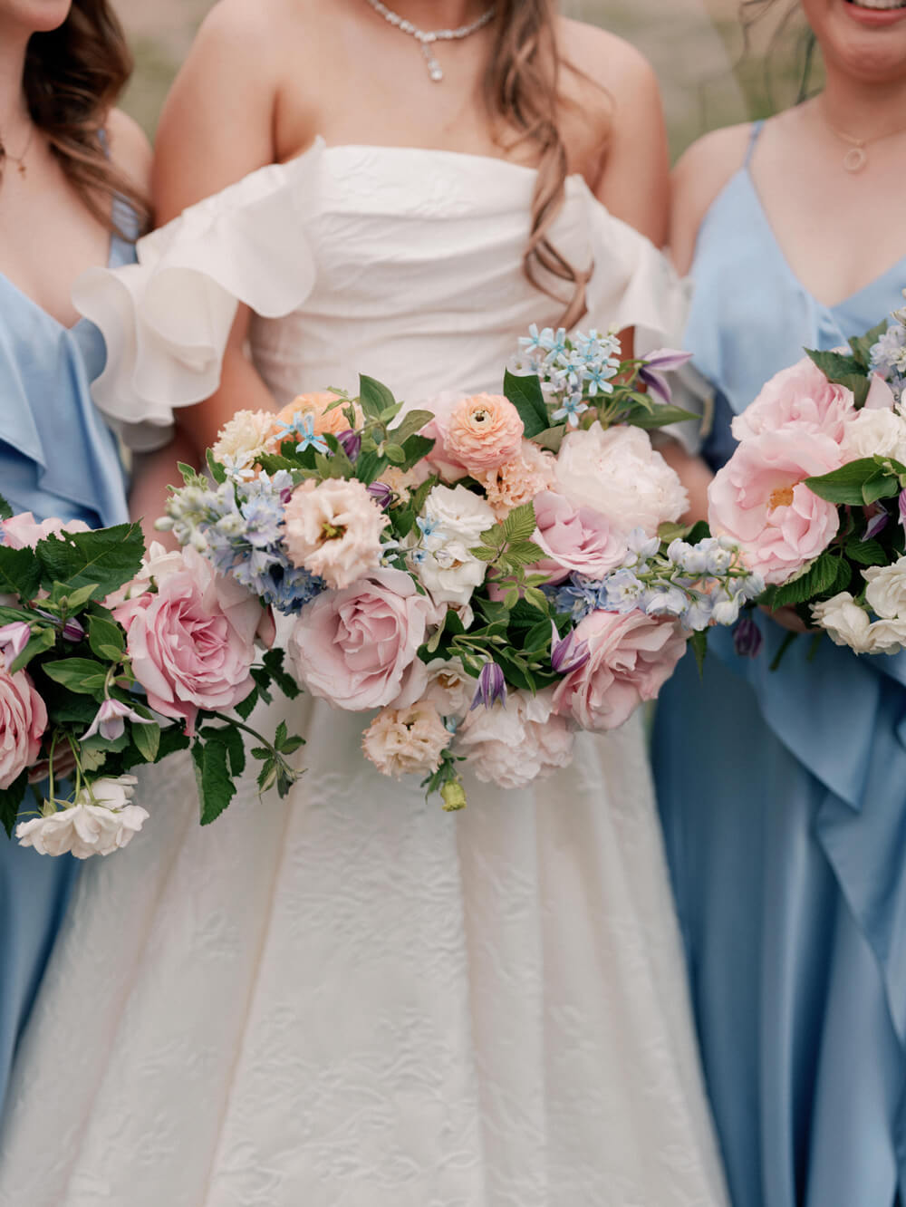 Bride in an off-the-shoulder wedding gown standing with bridesmaids in pastel blue dresses holding floral bouquets.