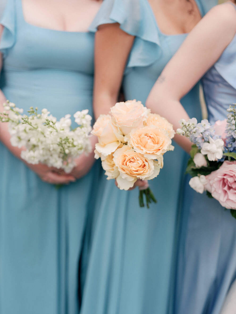Close-up of bridesmaids holding single-flower bouquets in soft peach and white tones during a summer wedding.