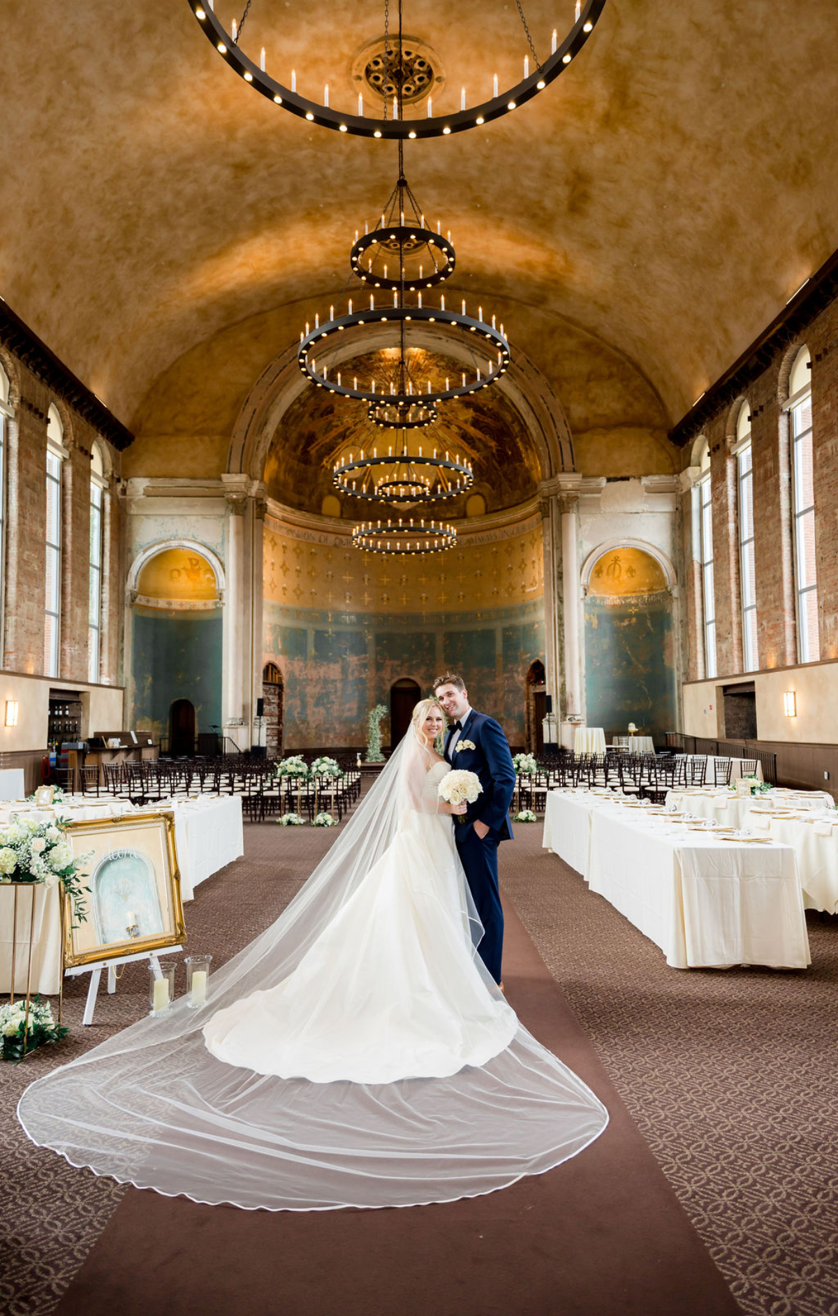 Choosing Your Wedding Invitation Style 4 Bride and groom stand arm in arm beneath ornate circular chandeliers in a grand, historic hall set for a wedding reception, with long tables, vaulted ceiling, and warm golden tones.