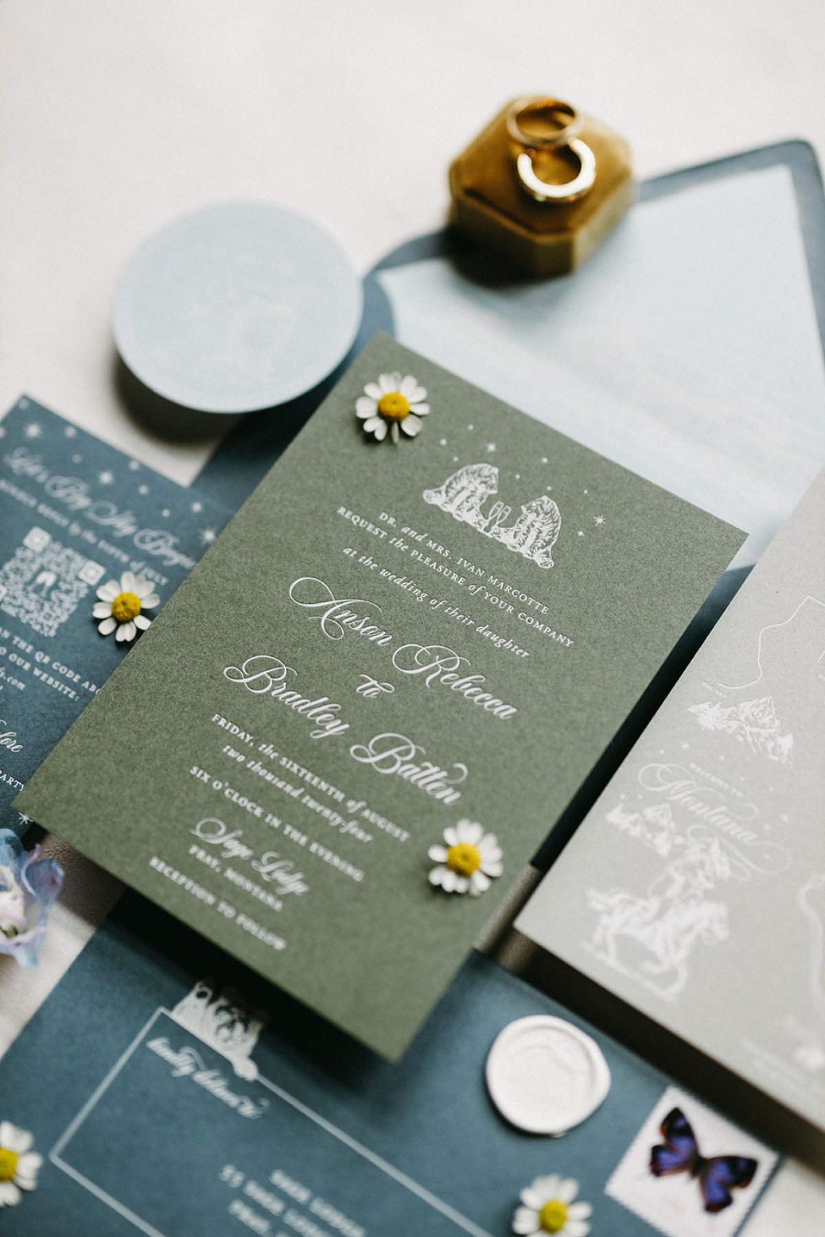 Flat lay of a Sage Lodge, Montana wedding invitation suite featuring sage green and blue stationery, custom bear illustration, western-inspired typography, wax seal, and day-of paper details.