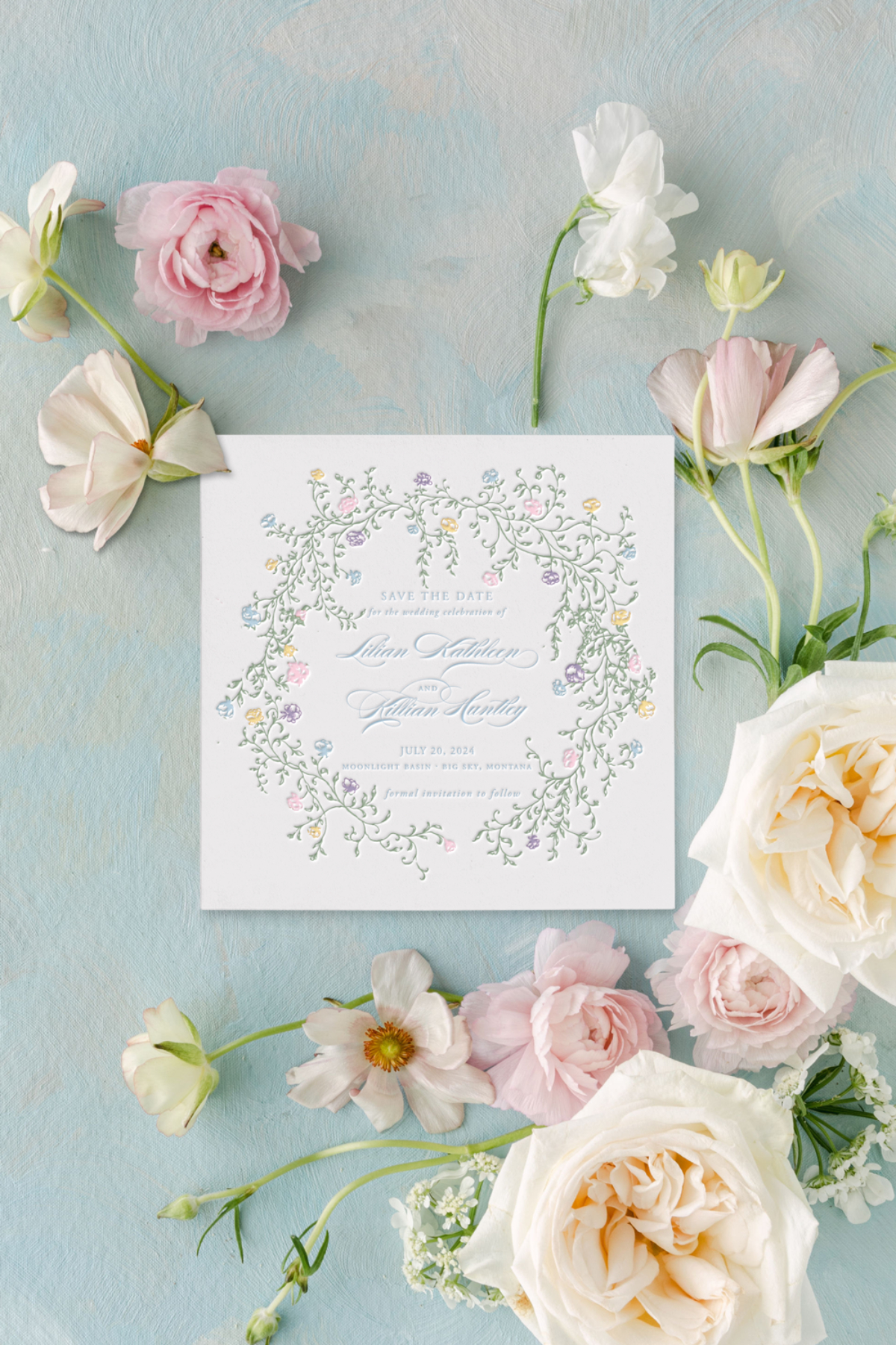 Flat lay of a floral save-the-date card surrounded by pastel roses and garden blooms on a soft blue textured background.
