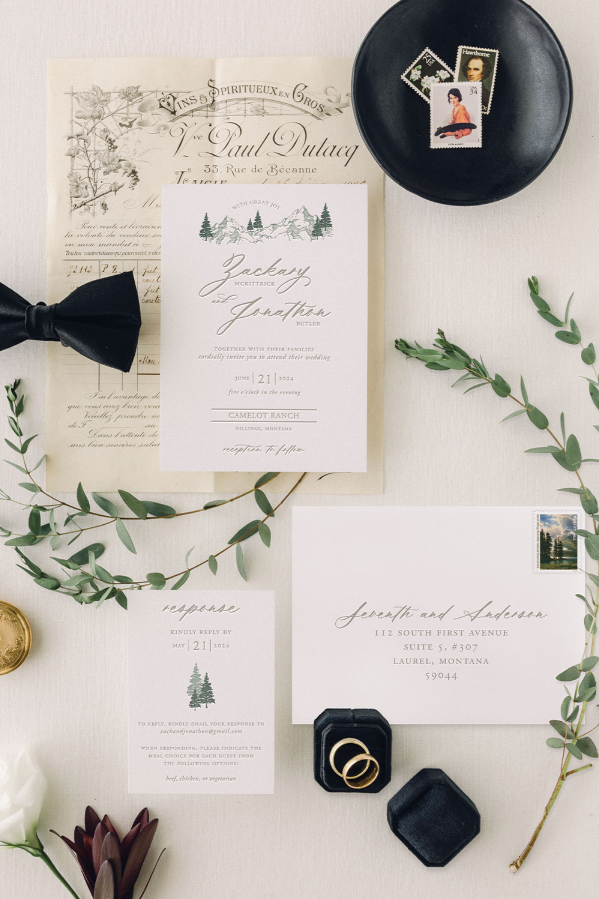Flat lay of a mountain-illustrated wedding invitation suite styled with vintage paper, stamps, greenery, a black bow tie, gold rings, and wax seal accessories. | wedding invitation questions