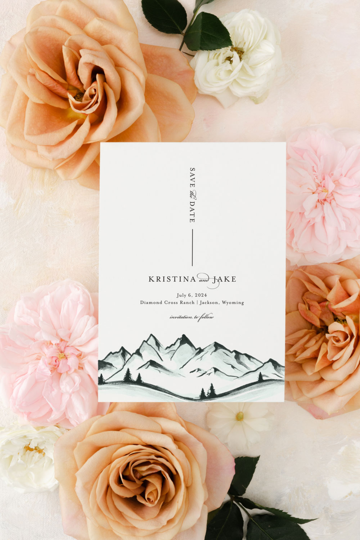 Flat lay of a minimalist mountain save-the-date card surrounded by peach and blush roses on a soft textured backdrop.