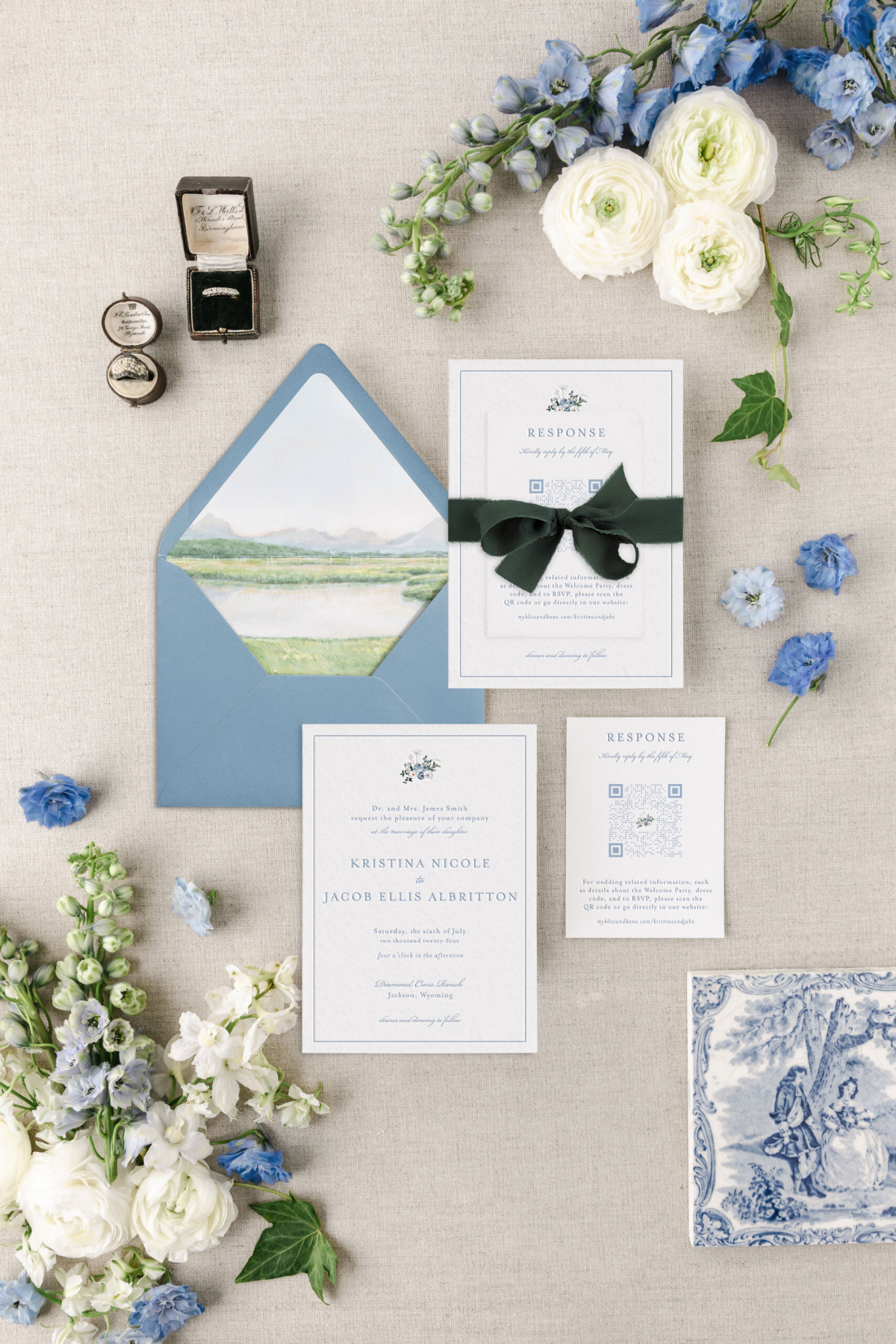 Choosing Your Wedding Invitation Style 10 Flat lay of blue envelope with painted landscape liner, response card tied with ribbon, vintage stamps, and flowers, reflecting a refined wedding invitatin style.