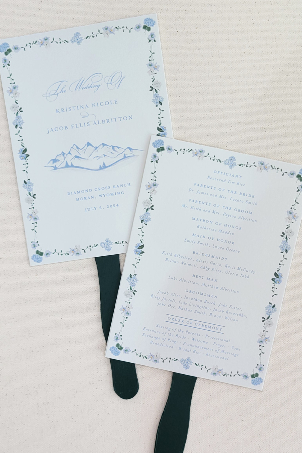 Choosing Your Wedding Invitation Style 9 Wedding ceremony program cards with blue floral borders and mountain motif, displayed on fabric fans, highlighting a classic, airy wedding invitatin style.