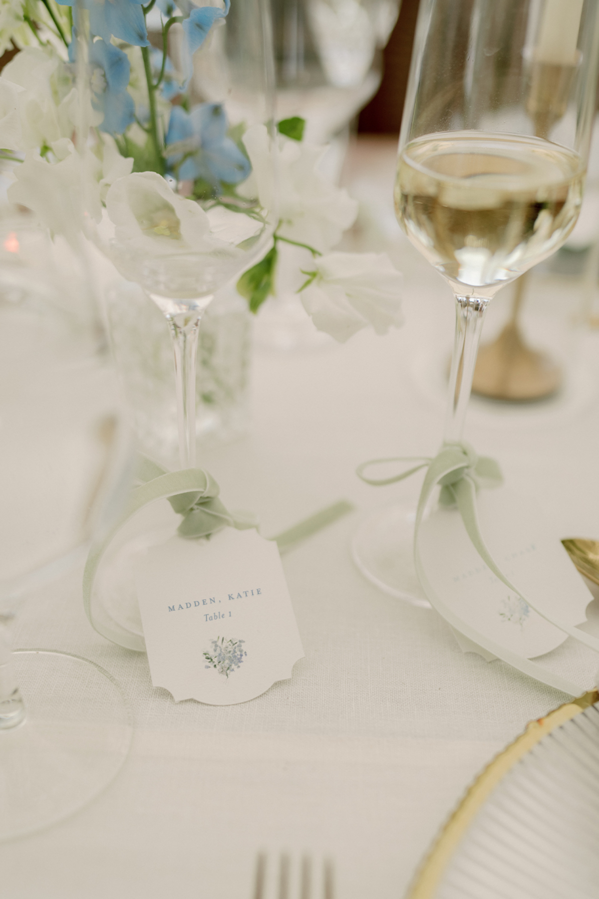Choosing Your Wedding Invitation Style 11 Place cards tied to champagne flutes with soft green ribbon on an elegant table setting, featuring delicate florals and a cohesive wedding invitatin style.