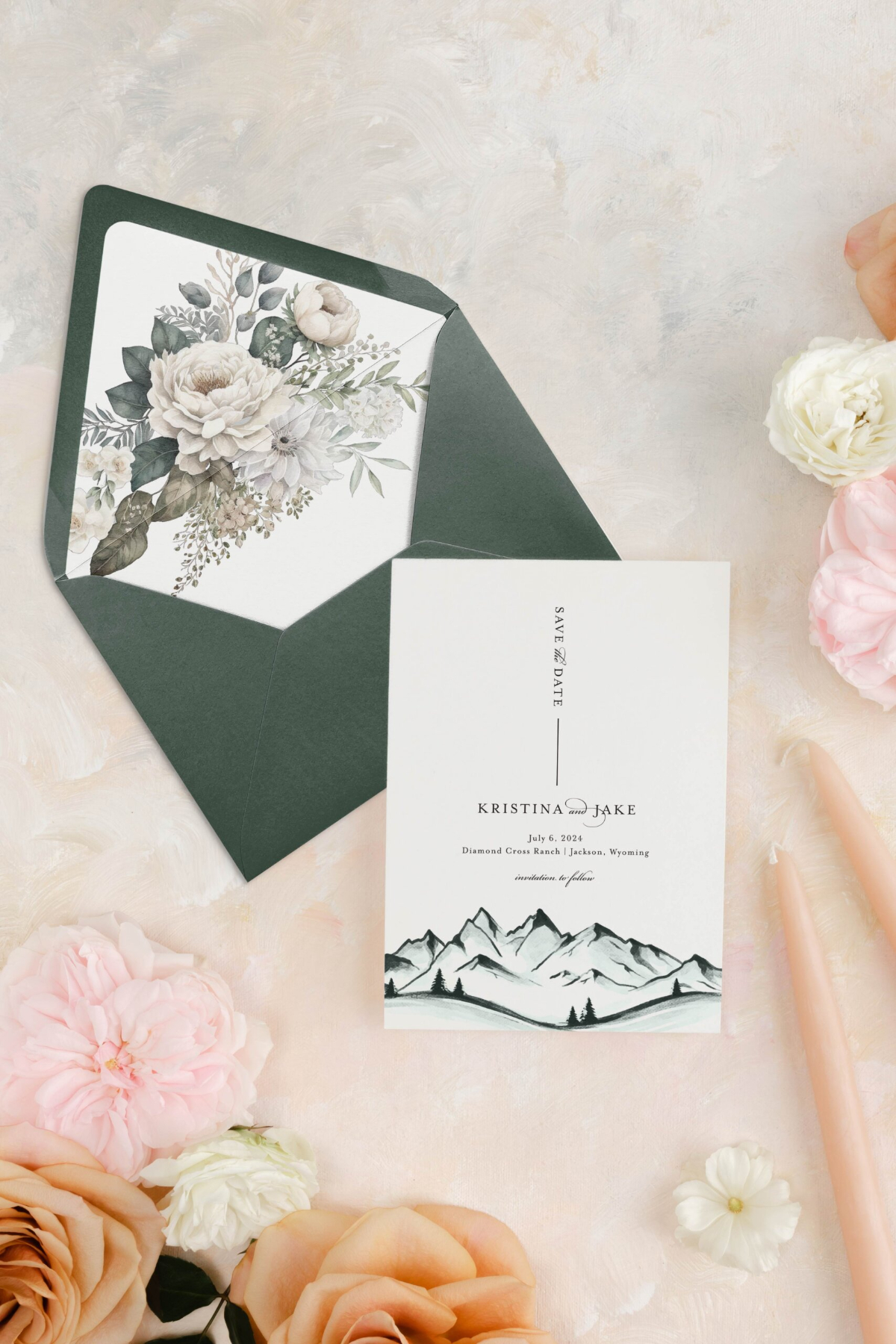Choosing Your Wedding Invitation Style 8 Save the date card with mountain illustration beside a dark green envelope with floral liner, styled with soft florals and candles, showcasing a romantic wedding invitatin style.