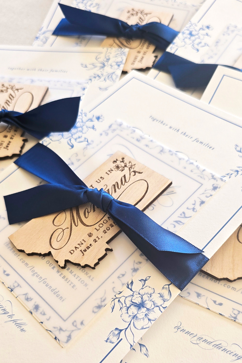 Guide to Luxury Wedding Invitations: What Makes Them Different & Why They Matter 4 Close-up of blue-and-white wedding invitation suites tied with navy silk ribbon and finished with custom wooden state-shaped tags. The layered cards, floral illustrations, and tactile details showcase the craftsmanship and intention behind luxury wedding invitations.