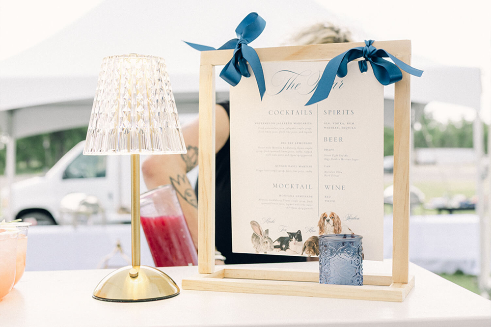Bar menu sign displayed on a wooden stand with silk ribbon ties, listing cocktails and spirits, thoughtfully designed to inform guests while adding a personal, welcoming touch to the reception.